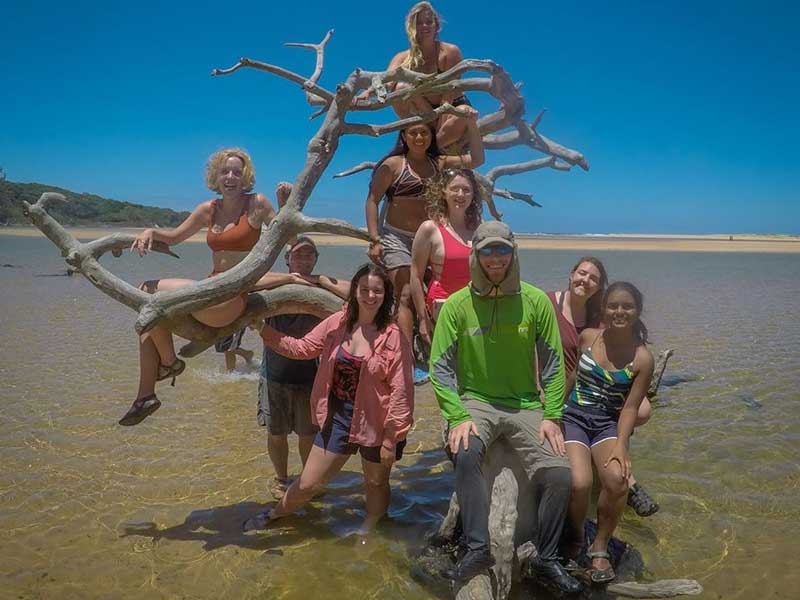 v.c.u. students pose on a piece of large driftwood on a beach