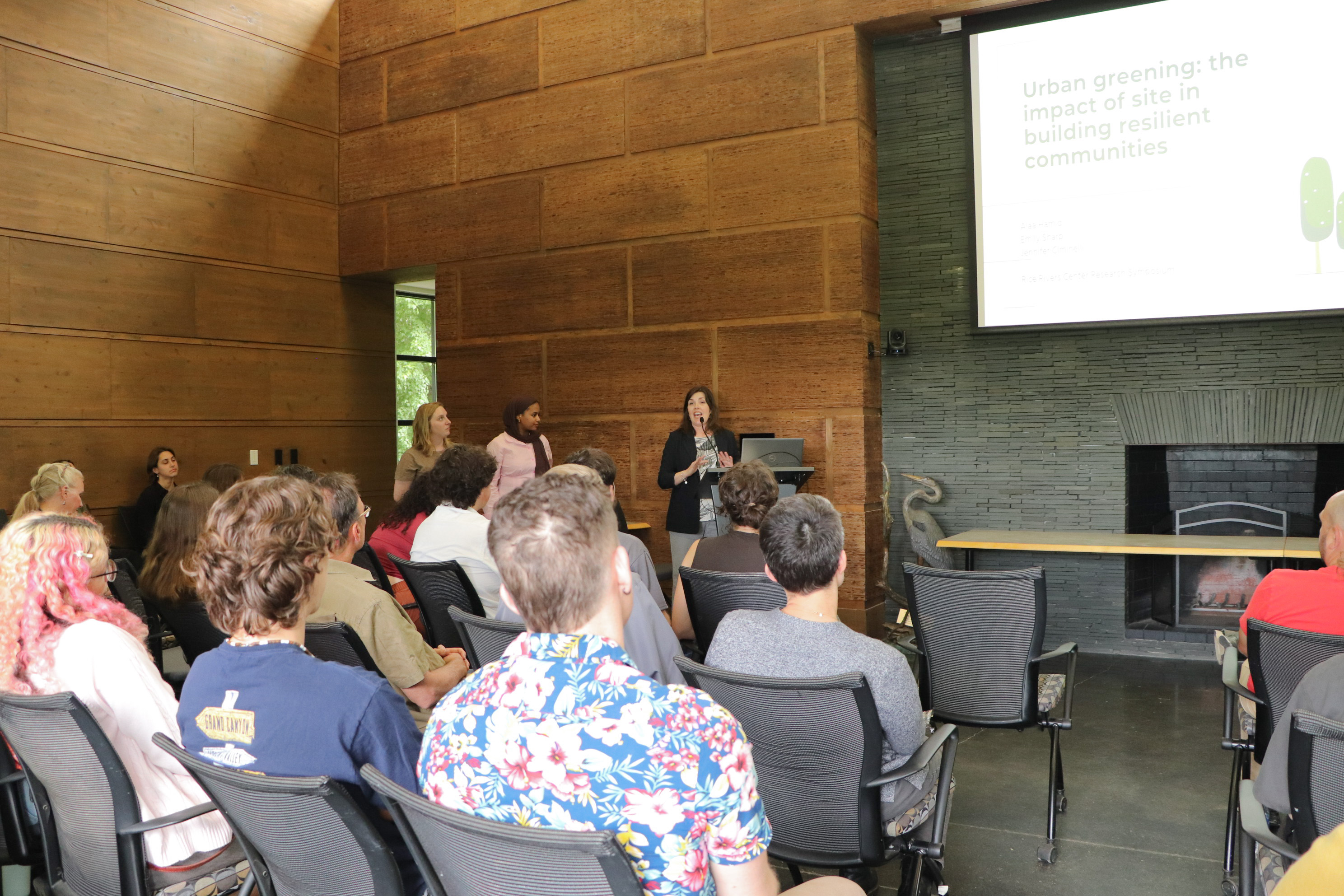 A group of students in chairs are watching presenters at a podium in front of a big screen on a wall
