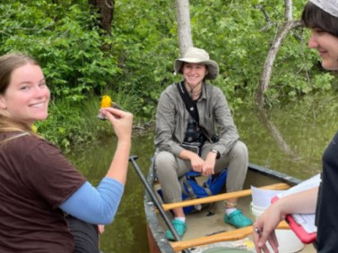 Three people sit on a canoe in the water, one holding a bird